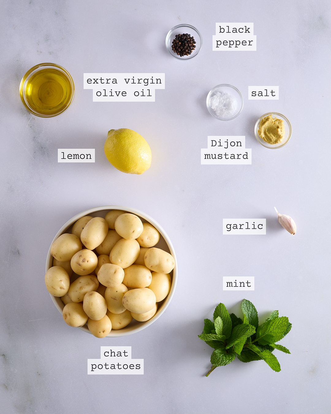 Flat lay of labelled ingredients for french-style potato salad (no mayo) with lemon & garlic on a light bench: chat potatoes, lemon, garlic, mint, extra virgin olive oil, Dijon mustard, sea salt, and black pepper.