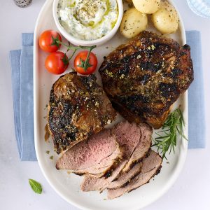 Overhead view of roasted lamb on a white platter with sliced lamb, baby potatoes, vine tomatoes and a bowl of cucumber yoghurt sauce, set on a pale bench with a blue napkin and rosemary sprigs.