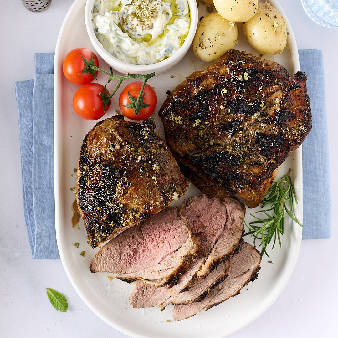 Overhead view of roasted lamb on a white platter with sliced lamb, baby potatoes, vine tomatoes and a bowl of cucumber yoghurt sauce, set on a pale bench with a blue napkin and rosemary sprigs.