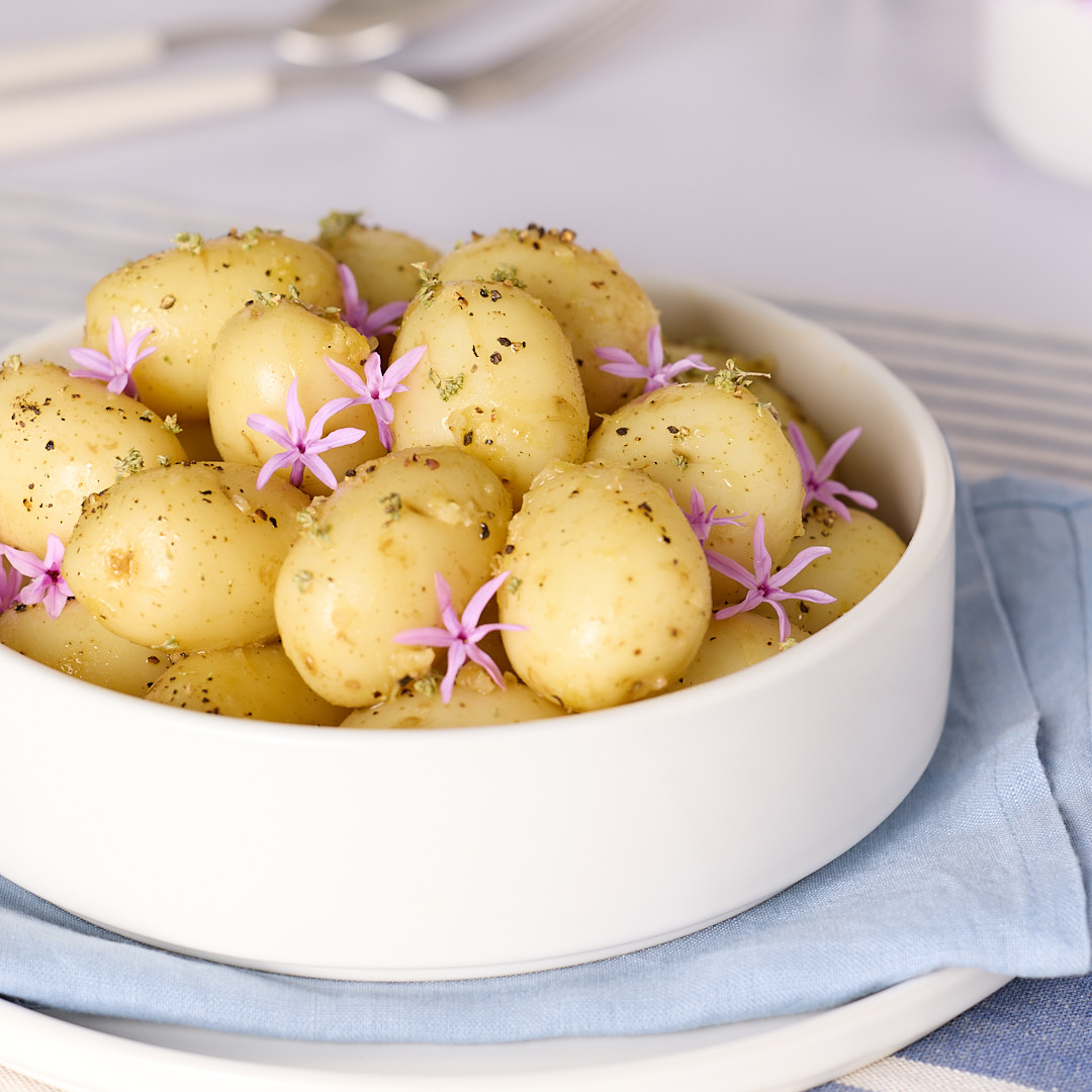 White ceramic bowl of baby potatoes tossed in a lemon and garlic vinaigrette, speckled with herbs and pepper, garnished with small purple flowers, sitting on a folded blue napkin on a striped tea towel on a bench.
