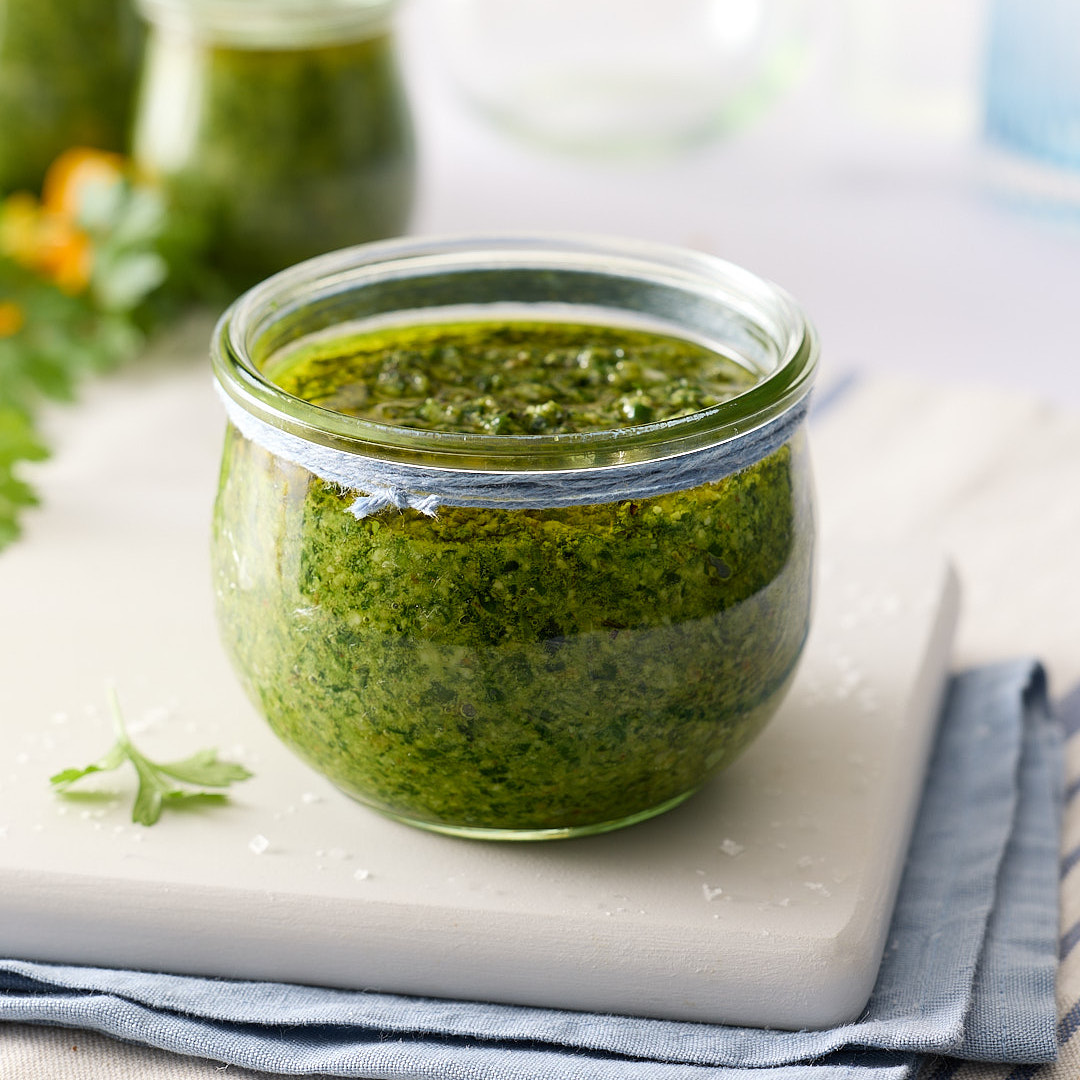 Jar of mixed herb walnut pesto topped with olive oil on a pale board and folded blue tea towel on a light bench, with extra jars blurred in the background.