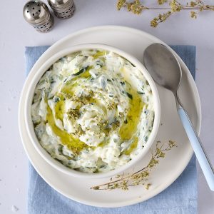 Creamy Greek tzatziki sauce in a white bowl, swirled with extra virgin olive oil and herbs, styled on a white plate over a blue linen napkin on a pale bench with a spoon and salt and pepper shakers.