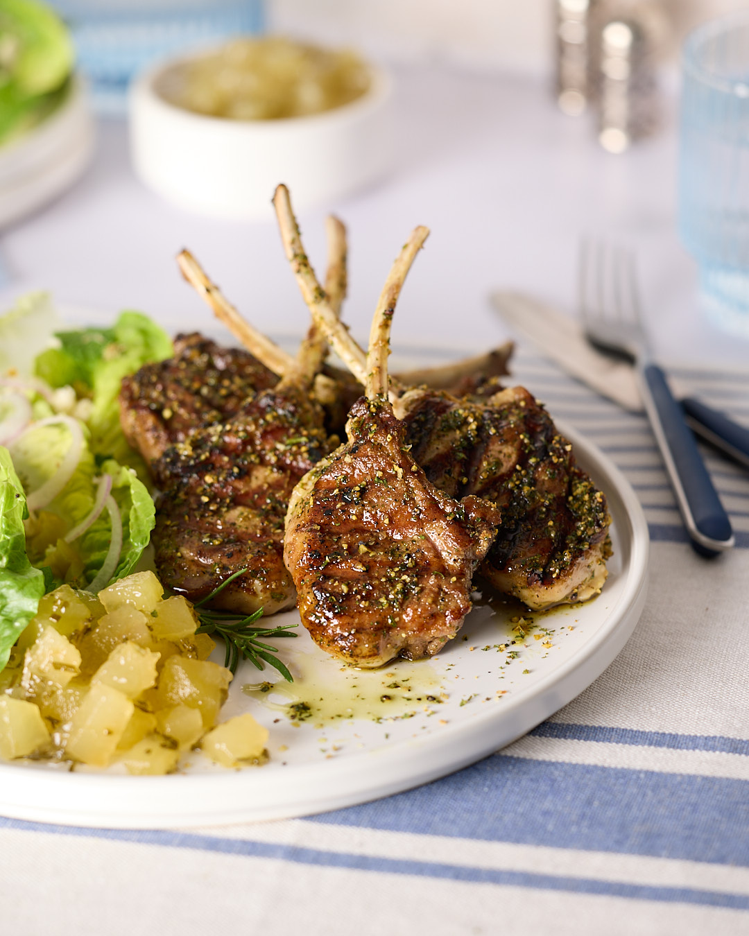 Pan-seared lamb chops on a plate with green salad and a spoonful of apple rosemary mint jam, with a small bowl of jam blurred in the background.