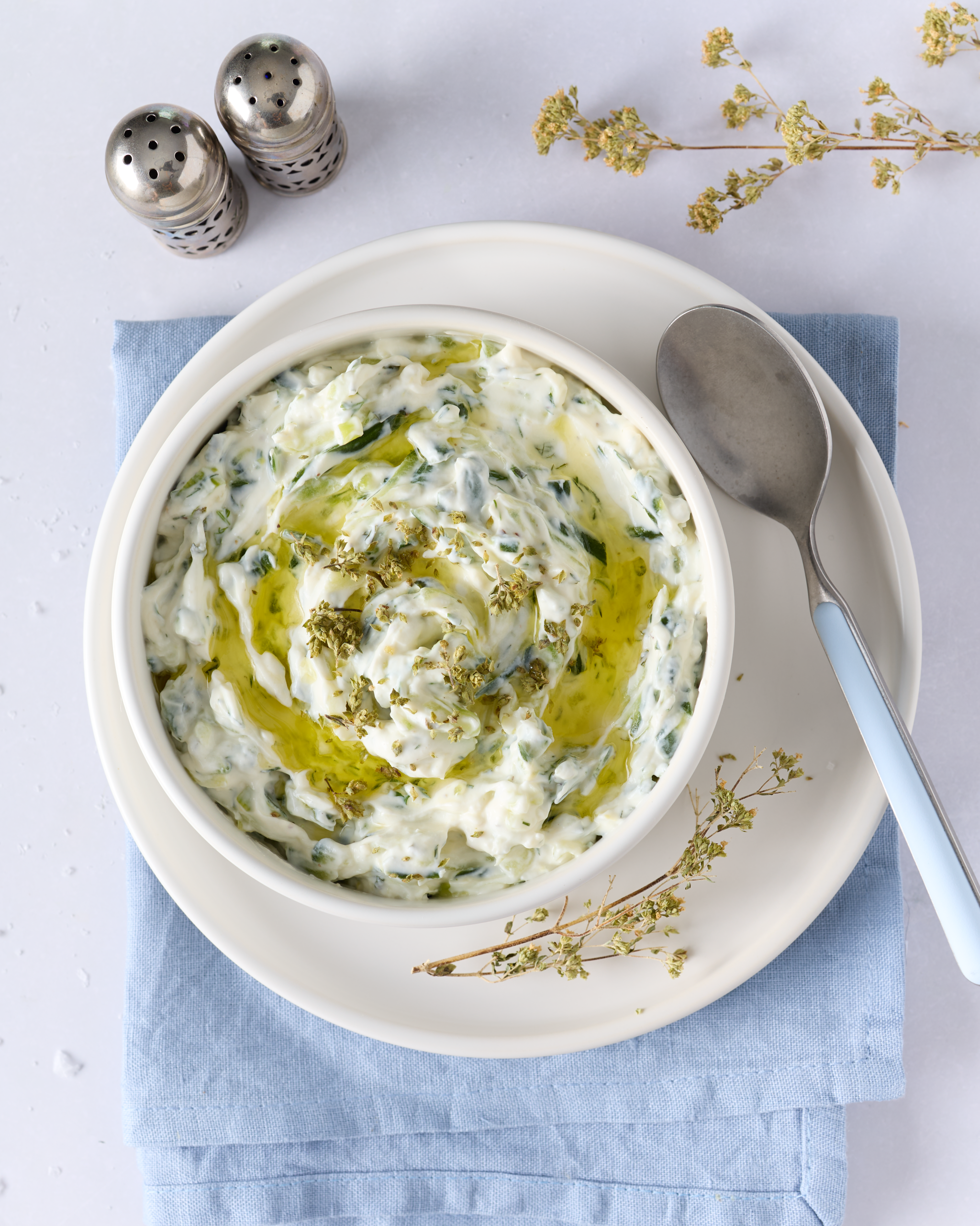 Overhead photo of a bowl of creamy tzatziki sauce for lamb swirled with olive oil and sprinkled with dried oregano, set on a white plate over a folded blue napkin, with a spoon and salt and pepper shakers nearby on a pale table.