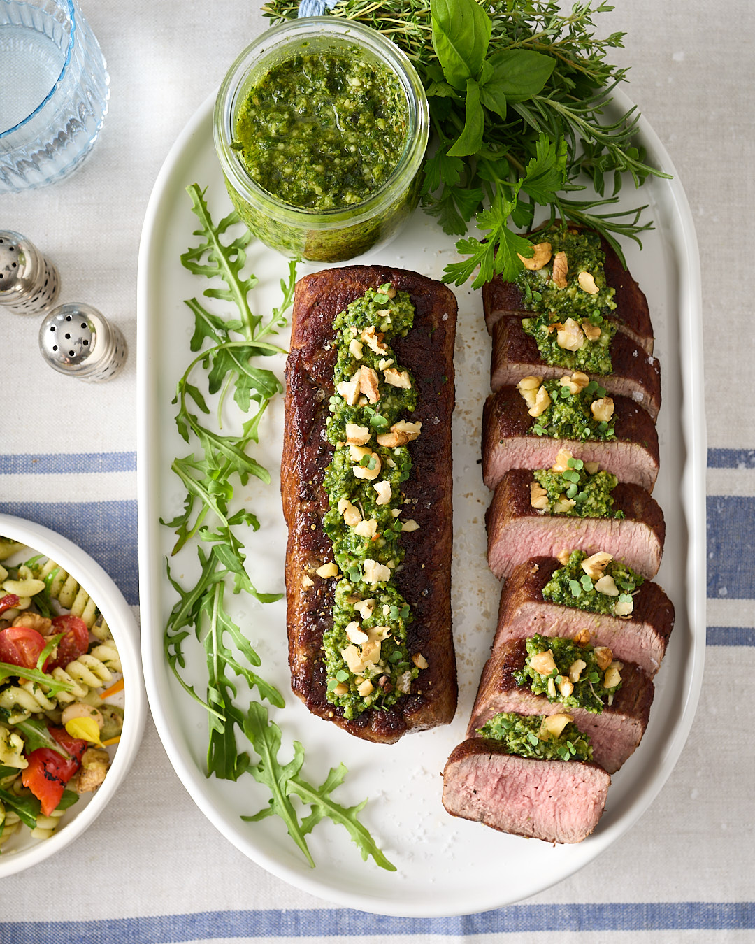 Sliced lamb roast and a whole lamb portion on a white serving platter, topped with spoonfuls of mixed herb walnut pesto and chopped walnuts, with a jar of pesto and fresh herb sprigs on the plate.