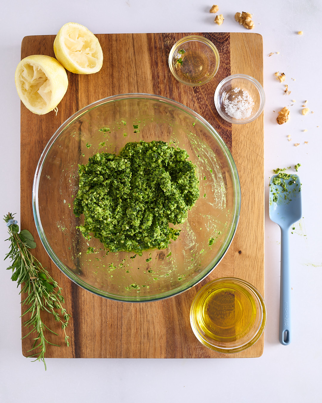 Glass mixing bowl of mixed herb walnut pesto on a wooden board, with squeezed lemon halves, olive oil, honey, sea salt, and a silicone spatula on a light bench.