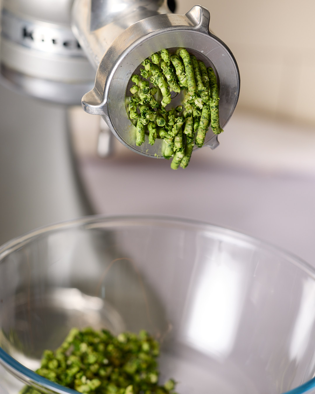 Close-up of a KitchenAid food grinder extruding strands of mixed herb walnut pesto into a glass bowl below.