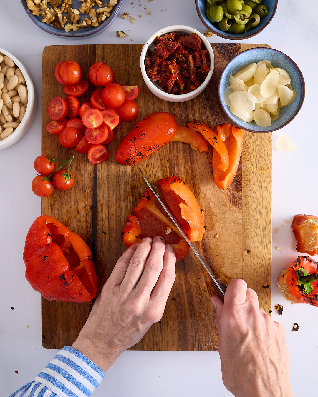 Sandra slicing a roasted red capsicum on a wooden chopping board, surrounded by prepped ingredients including cherry tomatoes, cannellini beans, semi sundried tomatoes, walnuts, Sicilian green olives, and pickled white onions.