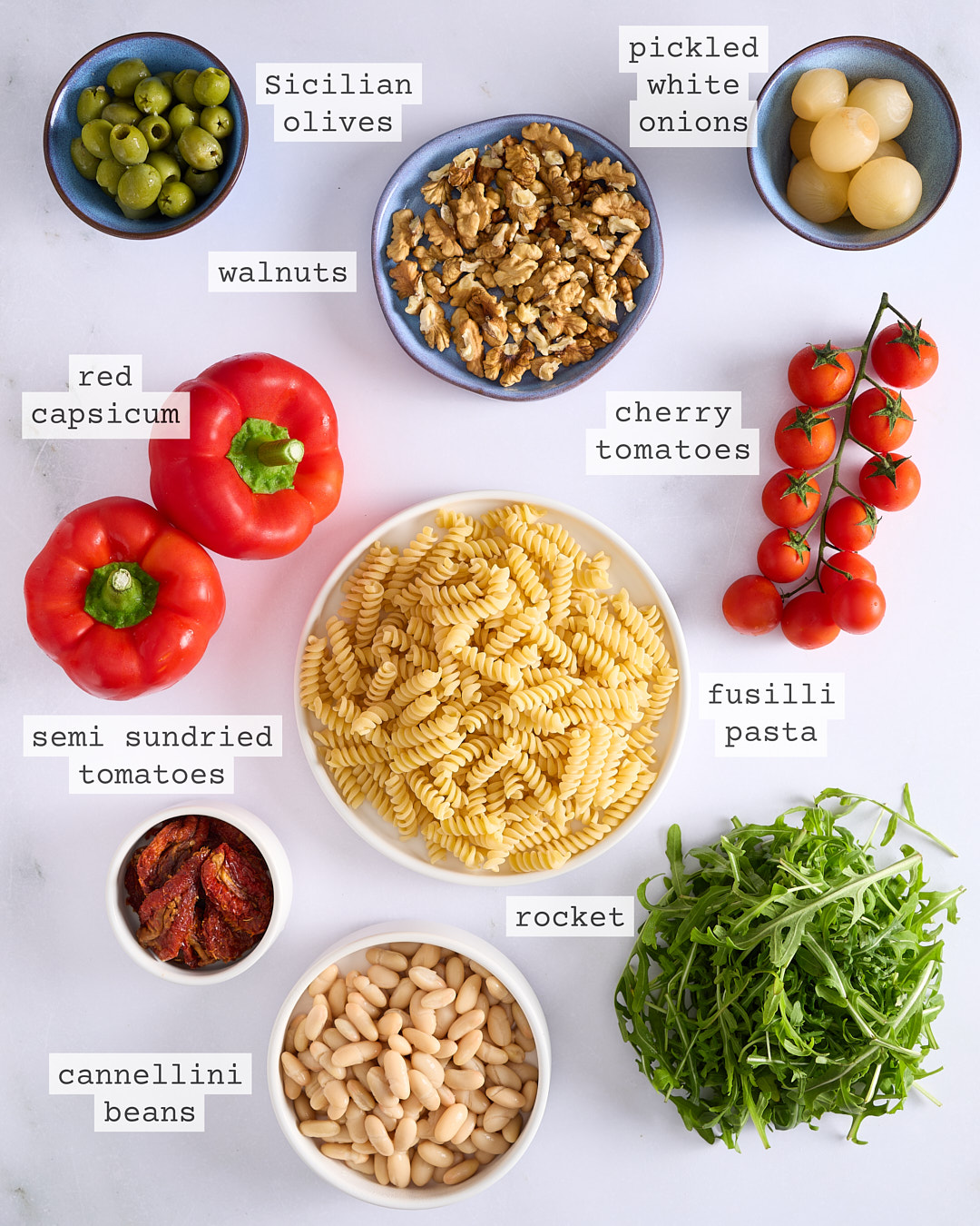 Overhead photo of labelled ingredients for a pesto pasta salad: fusilli pasta, red capsicum, cherry tomatoes, rocket, cannellini beans, semi sundried tomatoes, walnuts, Sicilian green olives, and pickled white onions arranged on a white surface.