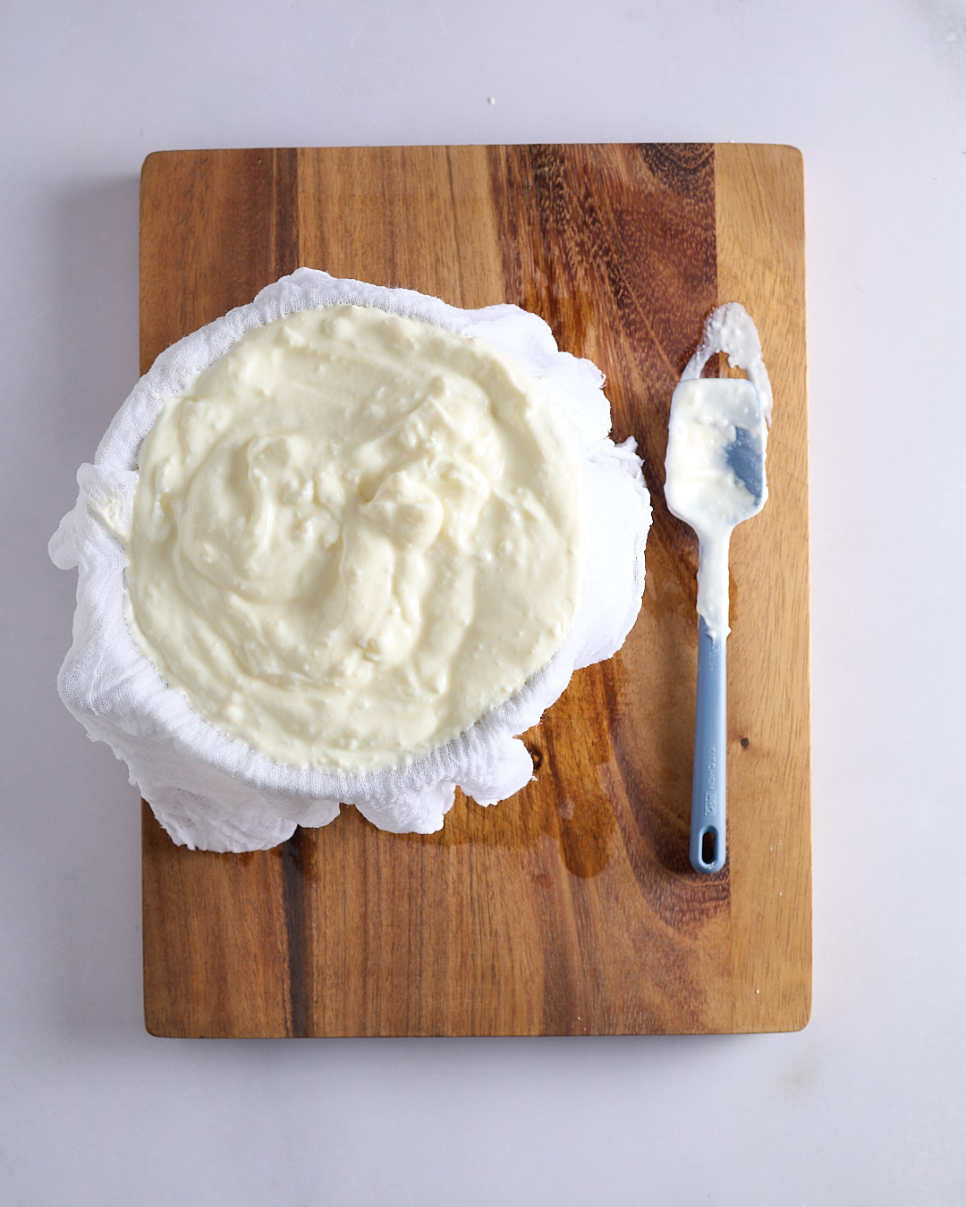 Thick Greek yoghurt spooned into a cheesecloth-lined sieve over a bowl on a wooden board on a pale bench, ready to be strained overnight.