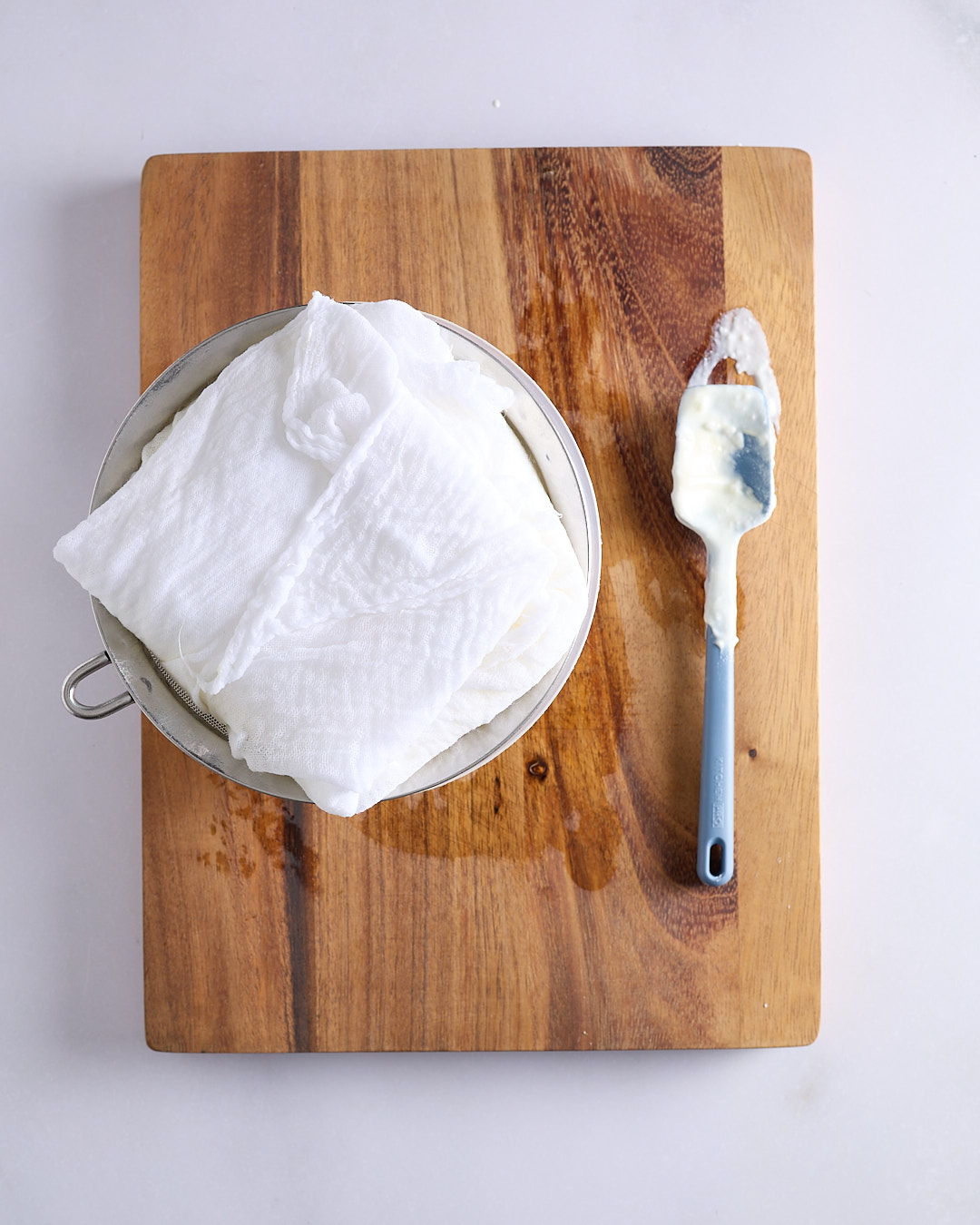 A large metal sieve lined with cheesecloth filled with Greek yoghurt and set over a bowl on a wooden board on a pale bench, with the yoghurt covered by overlapping cheesecloth and a blue silicone spatula alongside.