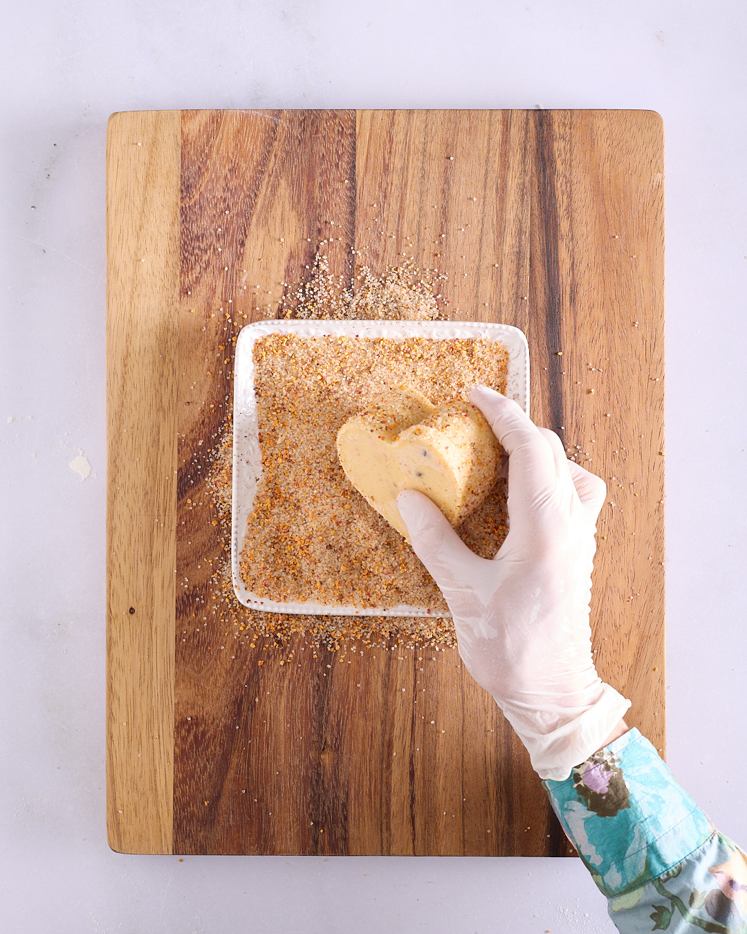 Sandra wearing a white plastic glove holding a stack of heart-shaped shortbread biscuits and rolling the edges through a square dish filled with citrus and pink peppercorn sugar.