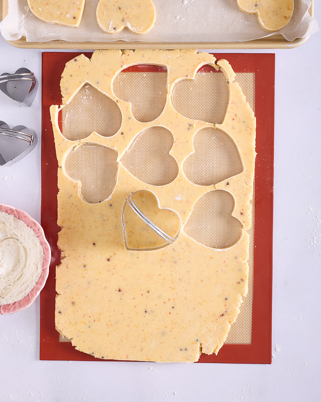Heart-shaped shortbread biscuits being cut from a sheet of dough on a silicone mat with a metal cookie cutter, with scraps and flour nearby.
