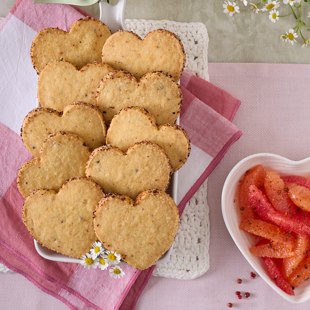 Heart-shaped pink peppercorn and grapefruit shortbread biscuits with a dark peppery sugar crust on the edges, served on a white platter next to fresh grapefruit segments and scattered pink peppercorns.