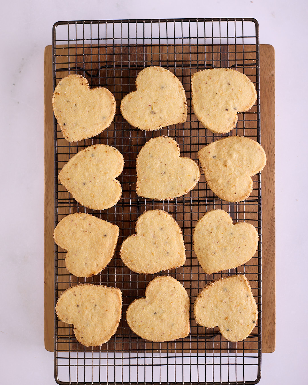 Twelve golden, heart-shaped pink peppercorn and grapefruit shortbread biscuits with a decorative citrus sugar crust on the edges, cooling on a black wire rack over a wooden board.
