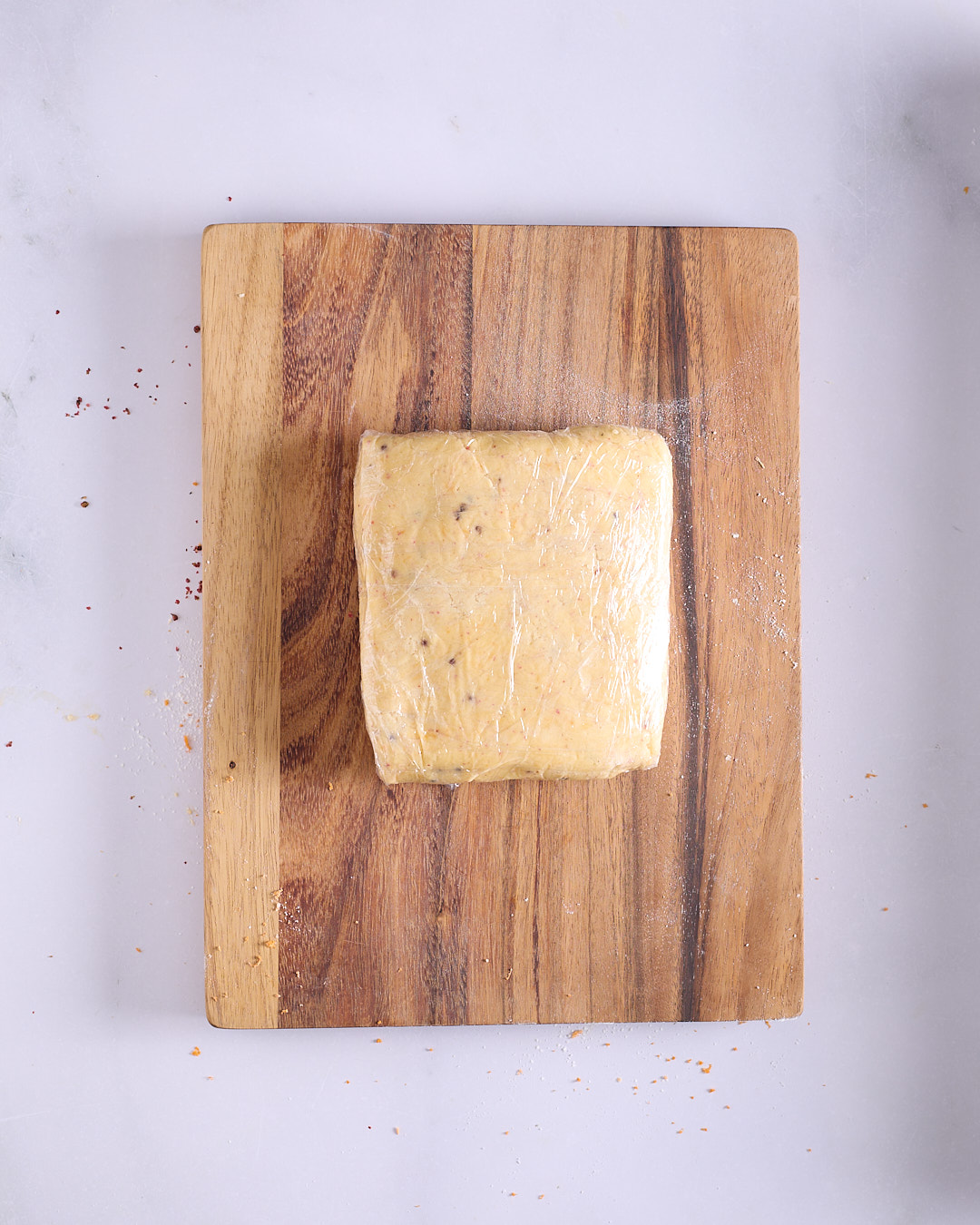 A thick rectangle of pink peppercorn shortbread dough wrapped tightly in clear plastic wrap on a wooden board.