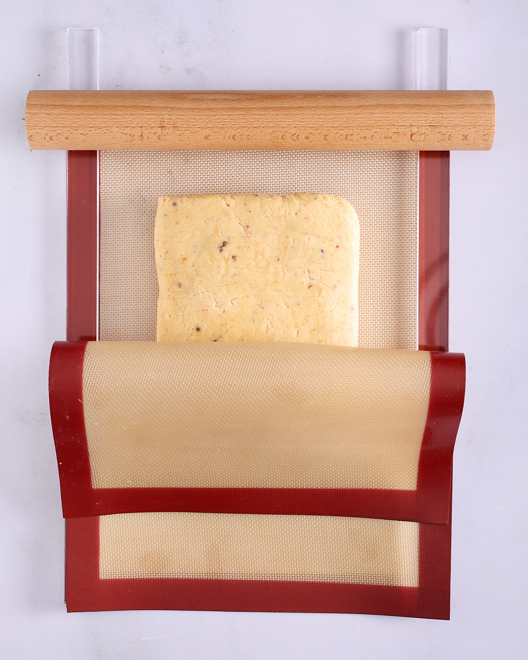 A firm slab of pink peppercorn shortbread dough positioned between two red-bordered silicone baking mats with clear acrylic rolling guides and a wooden rolling pin on top.