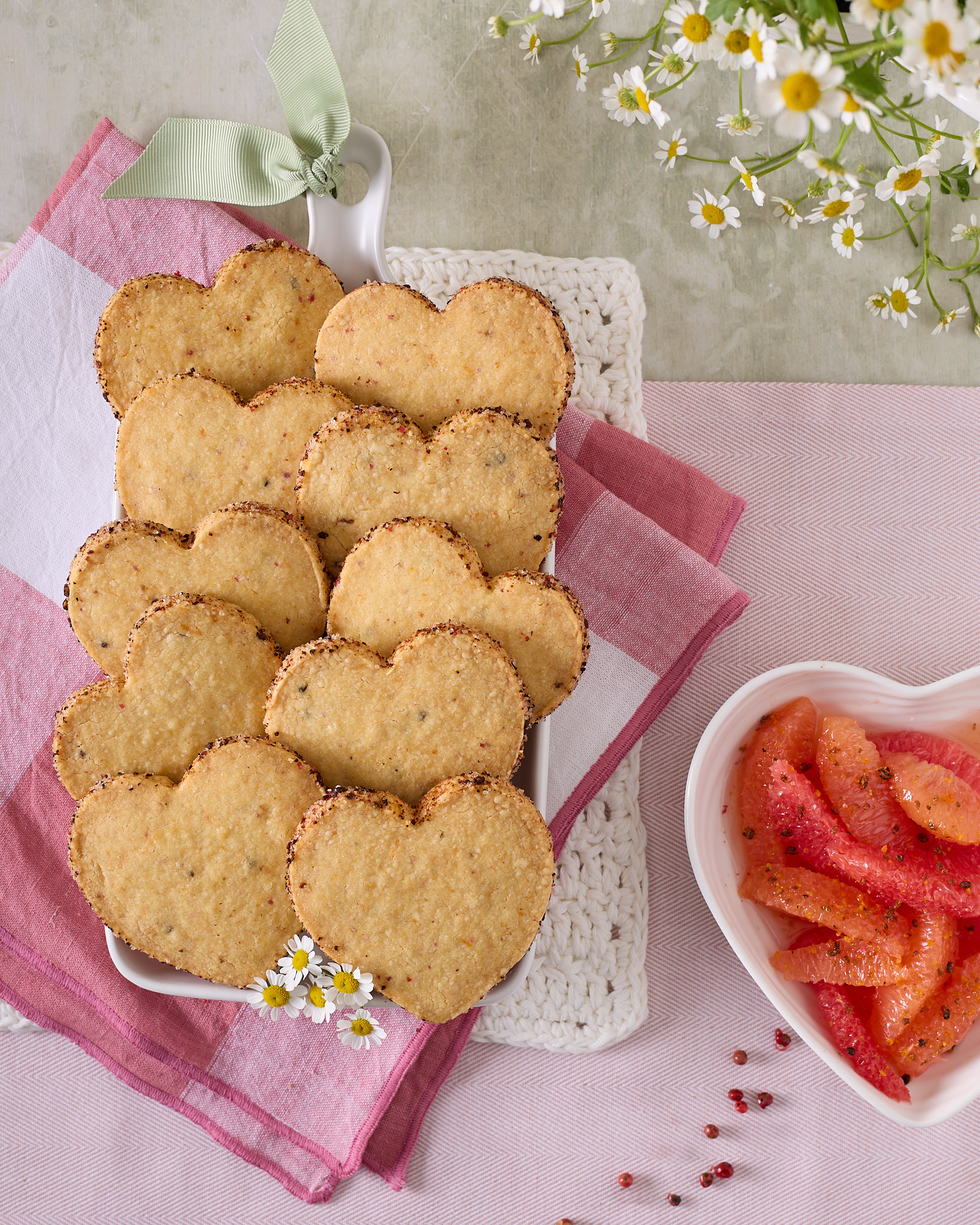 Heart-shaped pink peppercorn and grapefruit shortbread biscuits with a dark peppery sugar crust on the edges, served on a white platter next to fresh grapefruit segments and scattered pink peppercorns.
