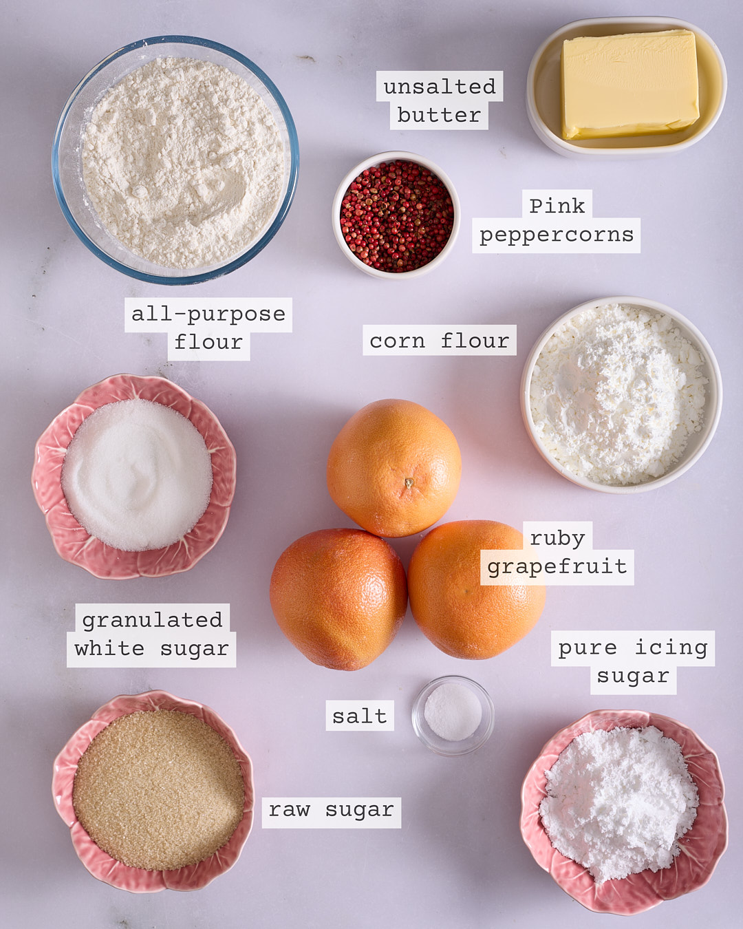 A flat lay of ingredients for pink peppercorn and grapefruit shortbread including unsalted butter, pink peppercorns, all-purpose flour, cornflour, ruby grapefruit, granulated white sugar, salt, raw sugar, and pure icing sugar, each clearly labelled.