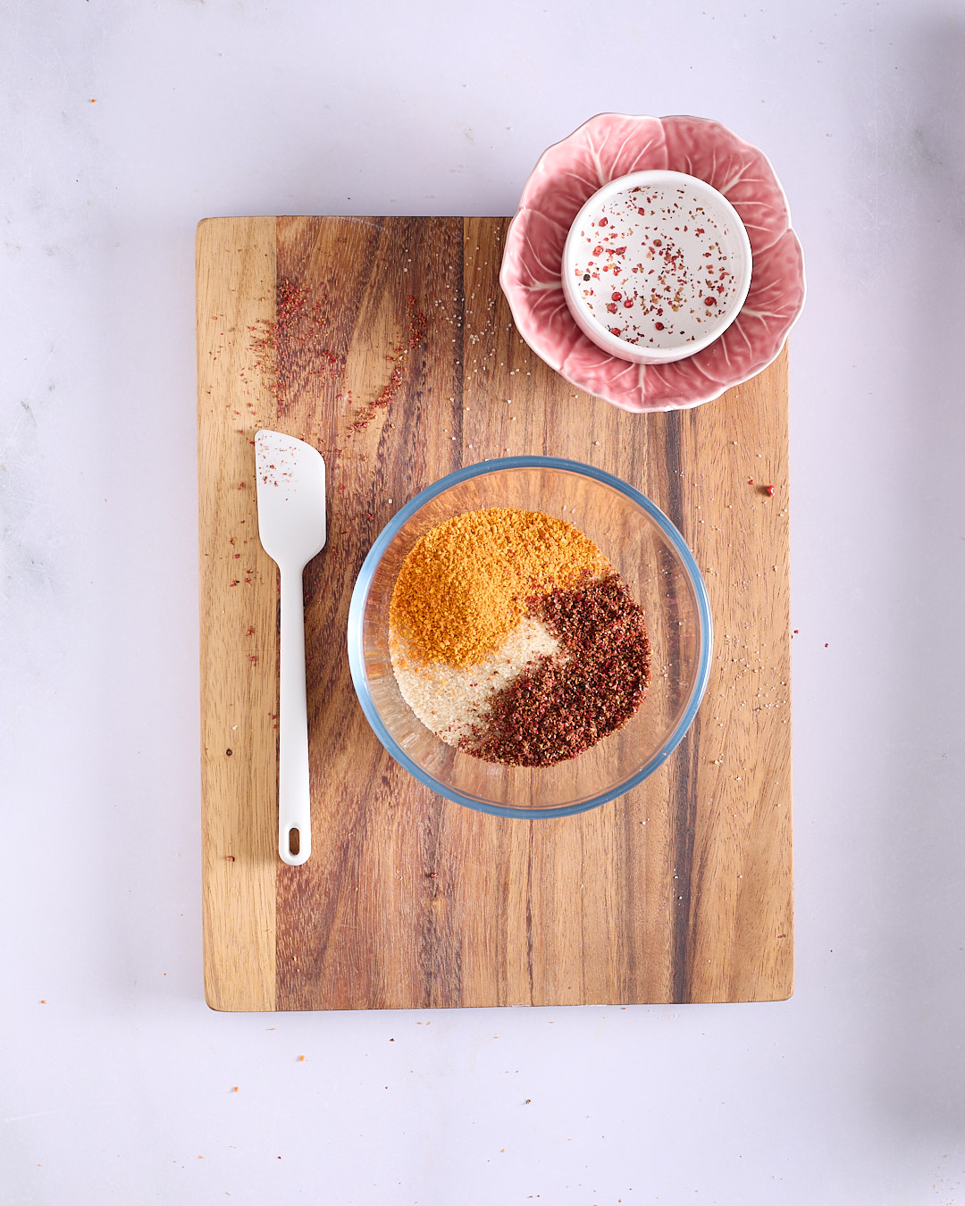 A glass bowl containing a mixture of granulated raw sugar, finely ground dehydrated grapefruit peel, and ground pink peppercorns, with a small white spatula ready for mixing.