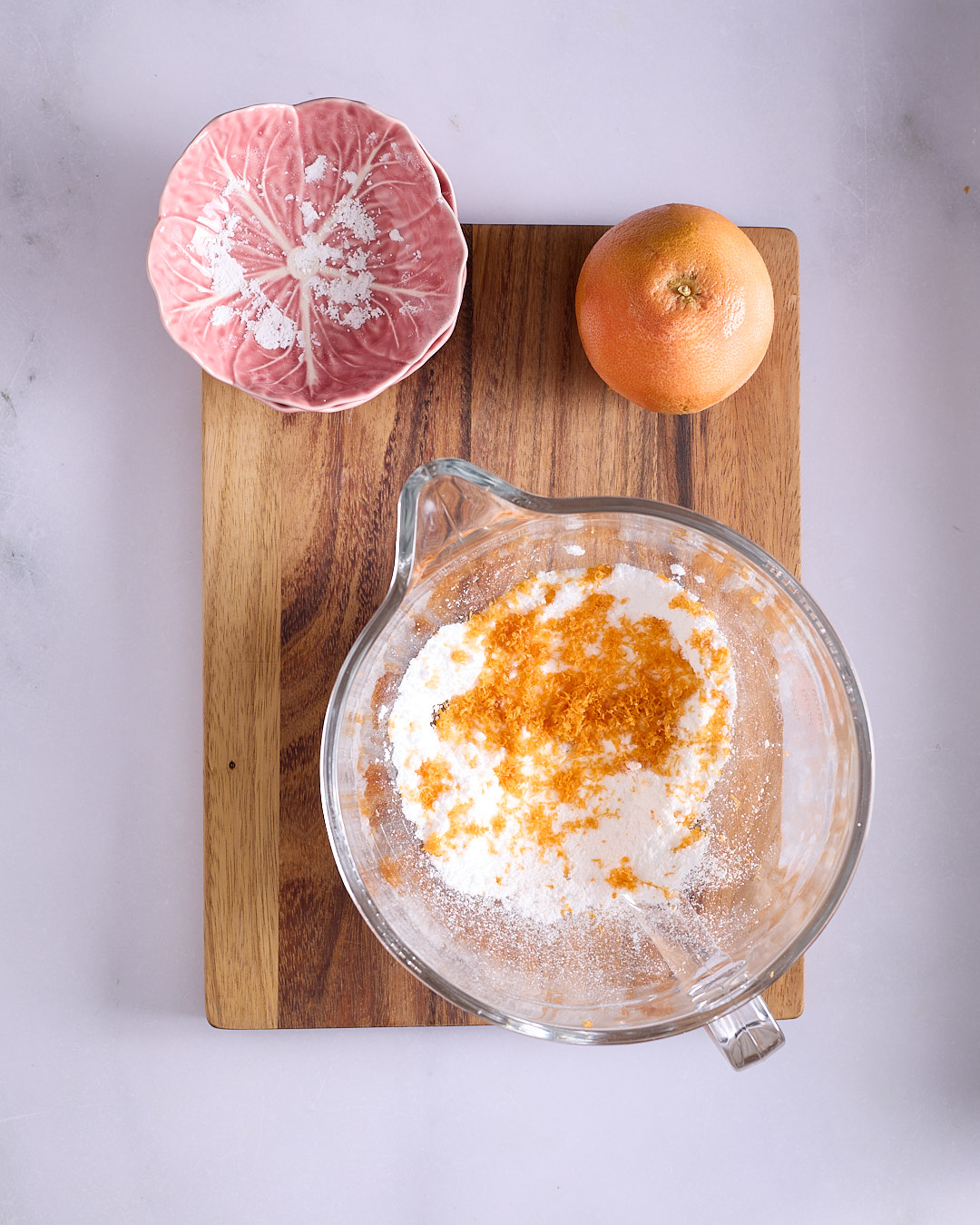 Freshly grated ruby grapefruit zest sitting on top of white sugar in a glass stand mixer bowl.