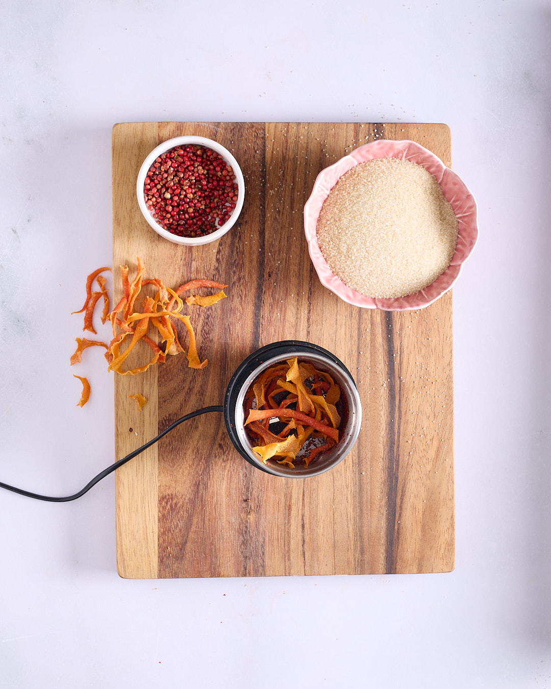 Strips of orange-ruby grapefruit peel being loaded into a black electric spice grinder on a wooden board, with raw sugar and whole pink peppercorns in bowls nearby.