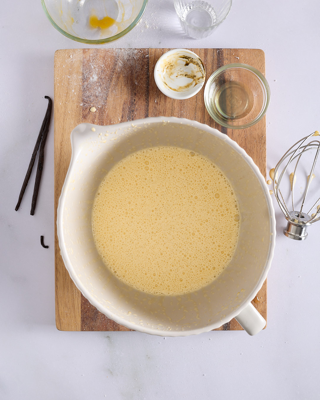 Pale yellow aerated egg yolk and vanilla mixture in a white mixing bowl with a whisk and vanilla bean pods nearby.