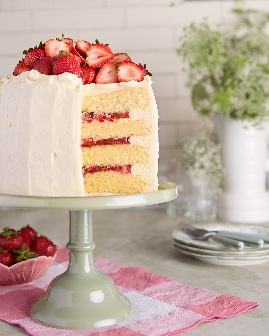 Tall strawberry basil white chocolate chiffon cake on a sage green cake stand with a slice removed, topped with fresh halved strawberries.