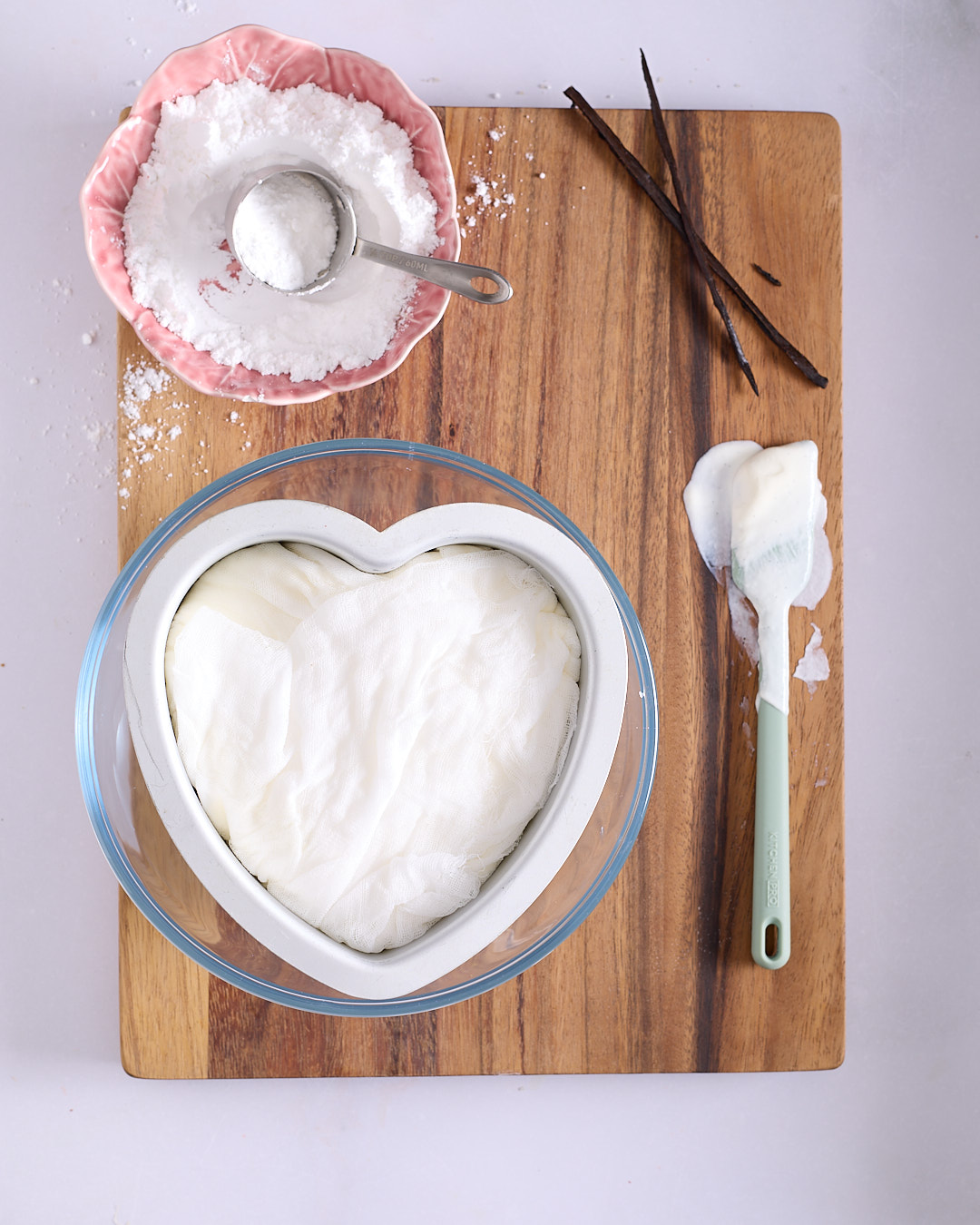 Heart shaped pan with white cheesecloth folded over the top of the yoghurt mixture, sitting inside a glass bowl.