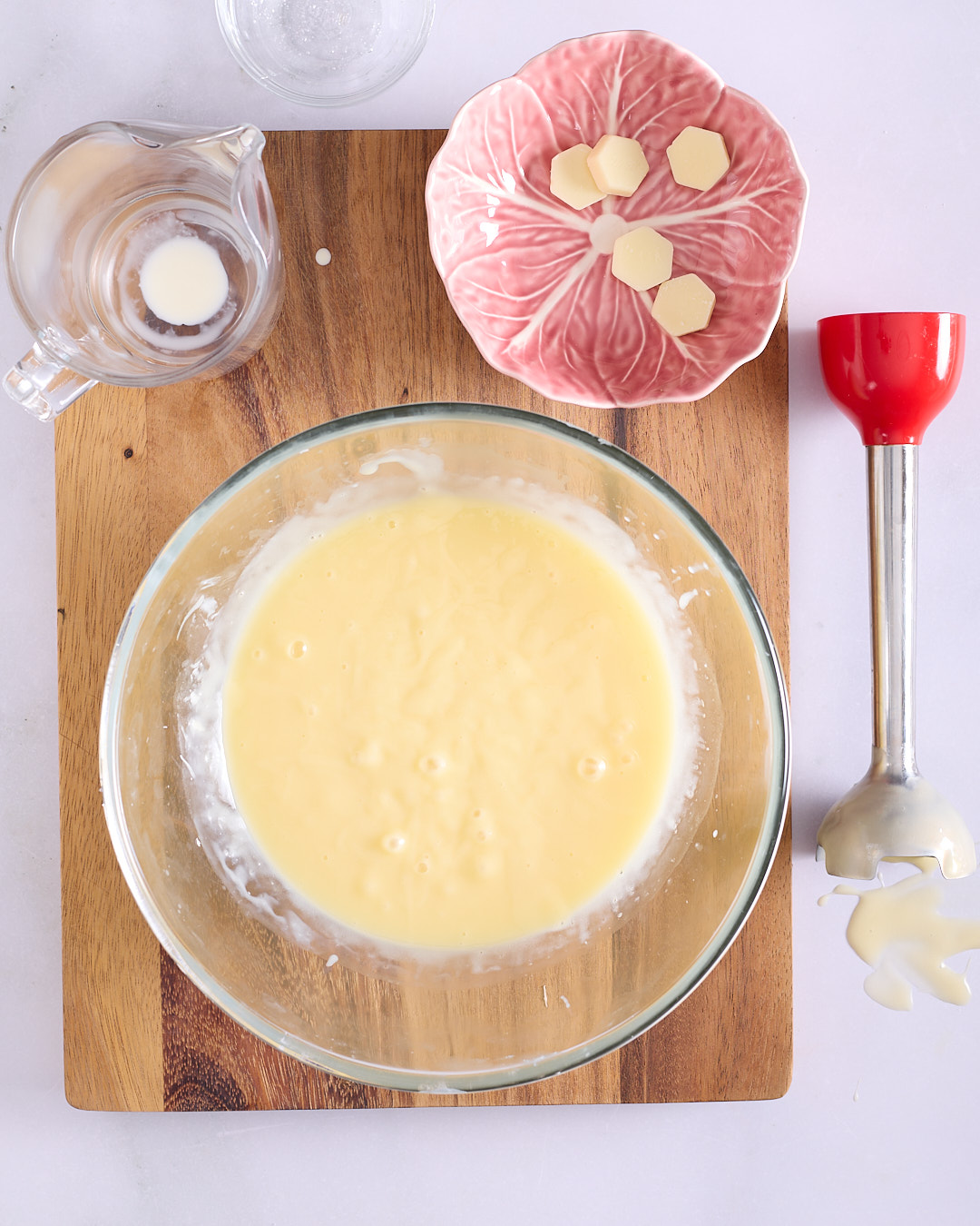Thick white chocolate ganache emulsion in a glass bowl with a red immersion blender and chocolate pieces on a pink dish.