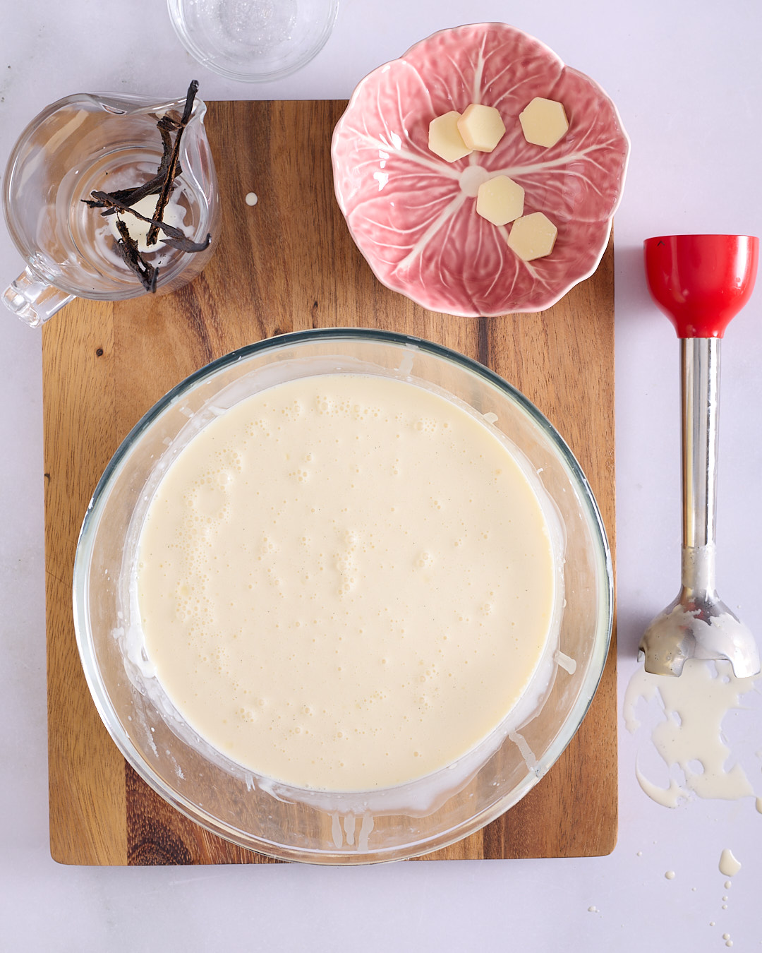 Large bowl of finished vanilla white chocolate ganache with vanilla bean hulls in a side jug and a red immersion blender.