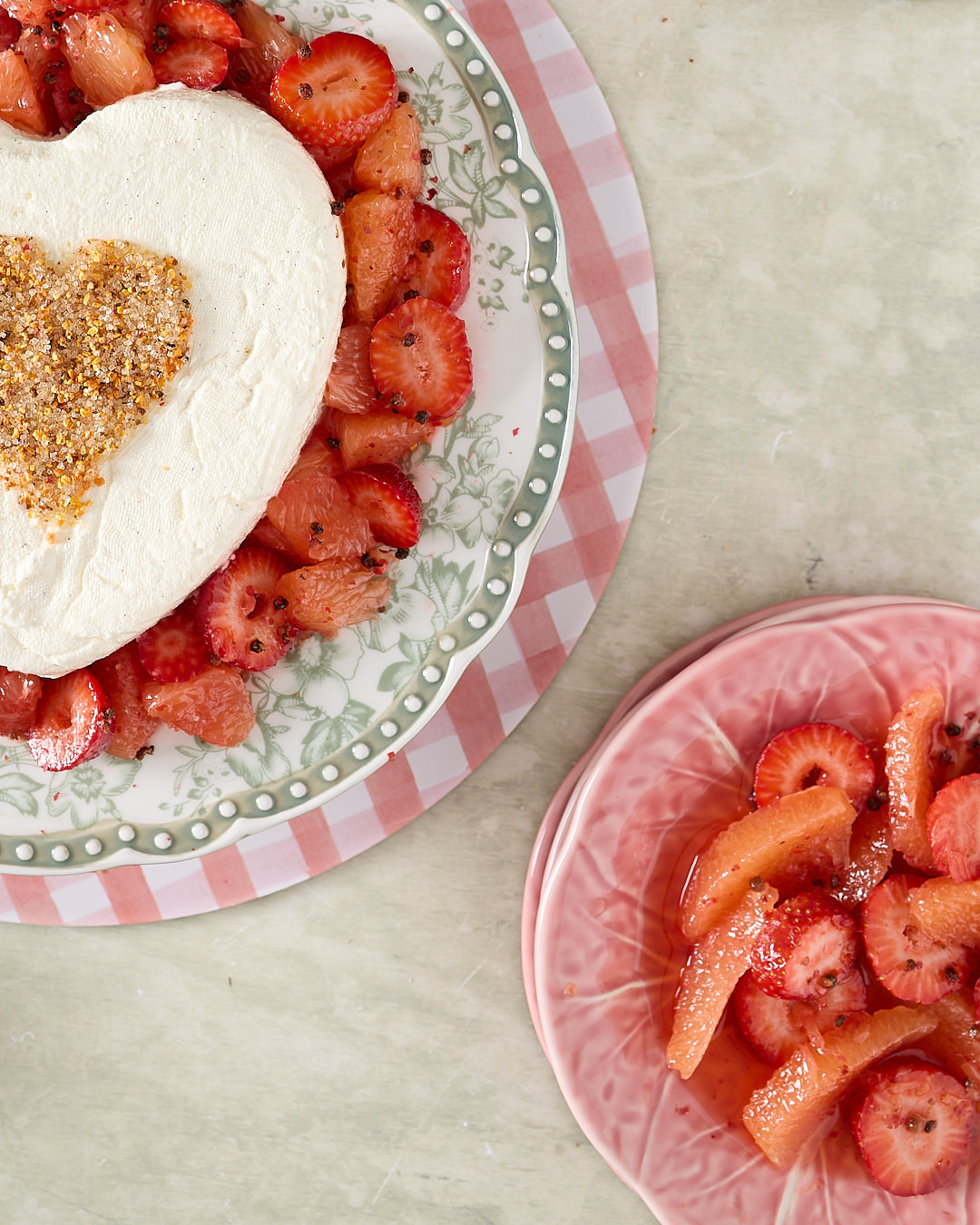 Close up of heart shaped yoghurt coeur à la crème on a decorative plate surrounded by grapefruit and strawberry compote with a stencilled heart of pink peppercorn sugar in the centre.