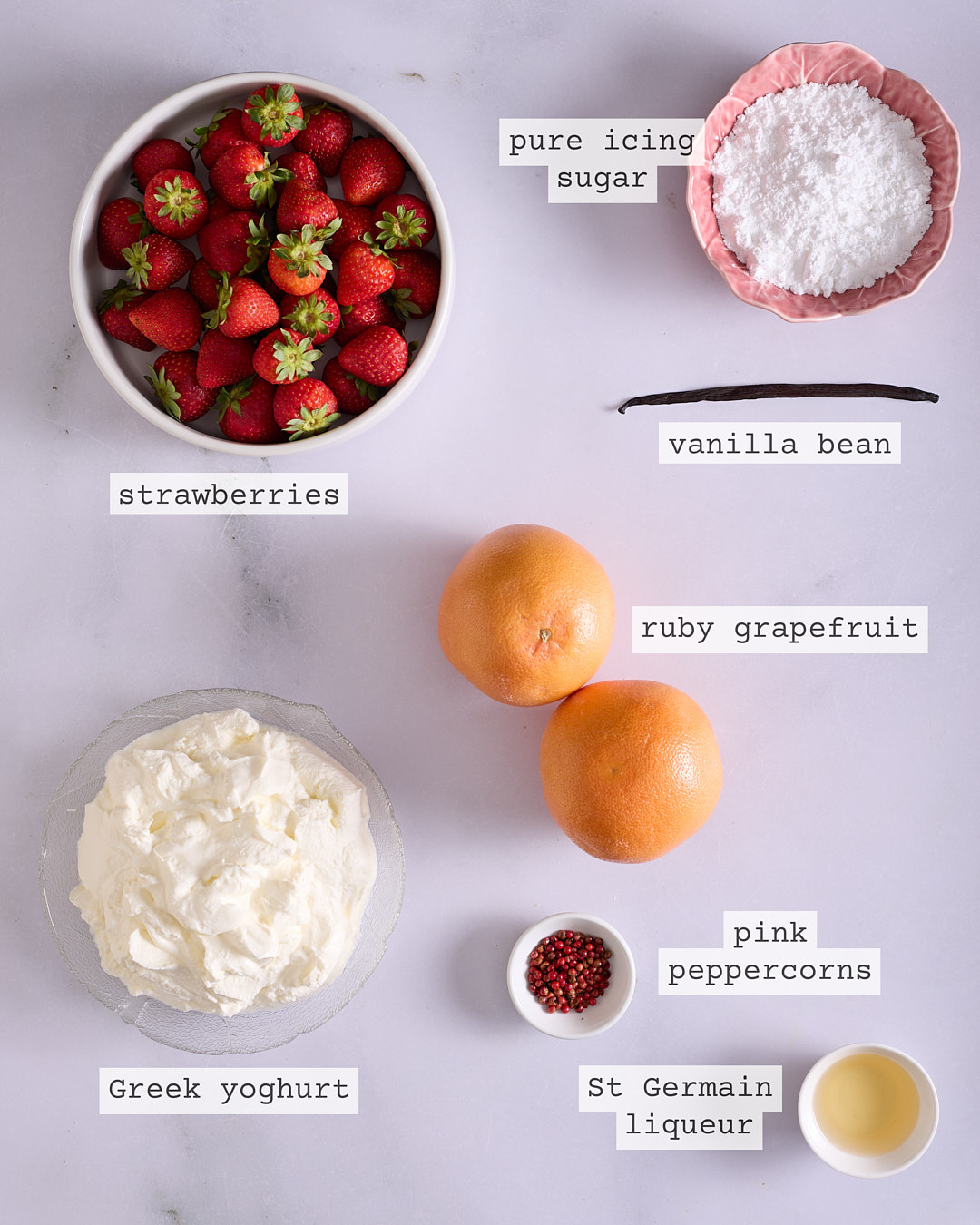 Flat lay of ingredients for yoghurt coeur à la crème including ruby grapefruit, strawberries, St Germain liqueur, pink peppercorns, Greek yoghurt, pure icing sugar, and a vanilla bean, each with a printed label.