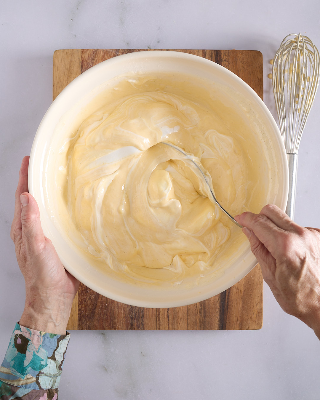 Sandra using a large metal spoon to gently fold white meringue into a pale-yellow cake batter in a large ceramic bowl with a whisk in the background.