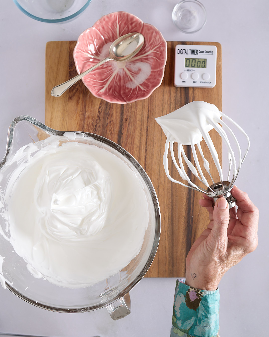 Sandra holding a whisk attachment with a stiff peak of glossy white meringue over a glass mixing bowl with a digital timer in the background.