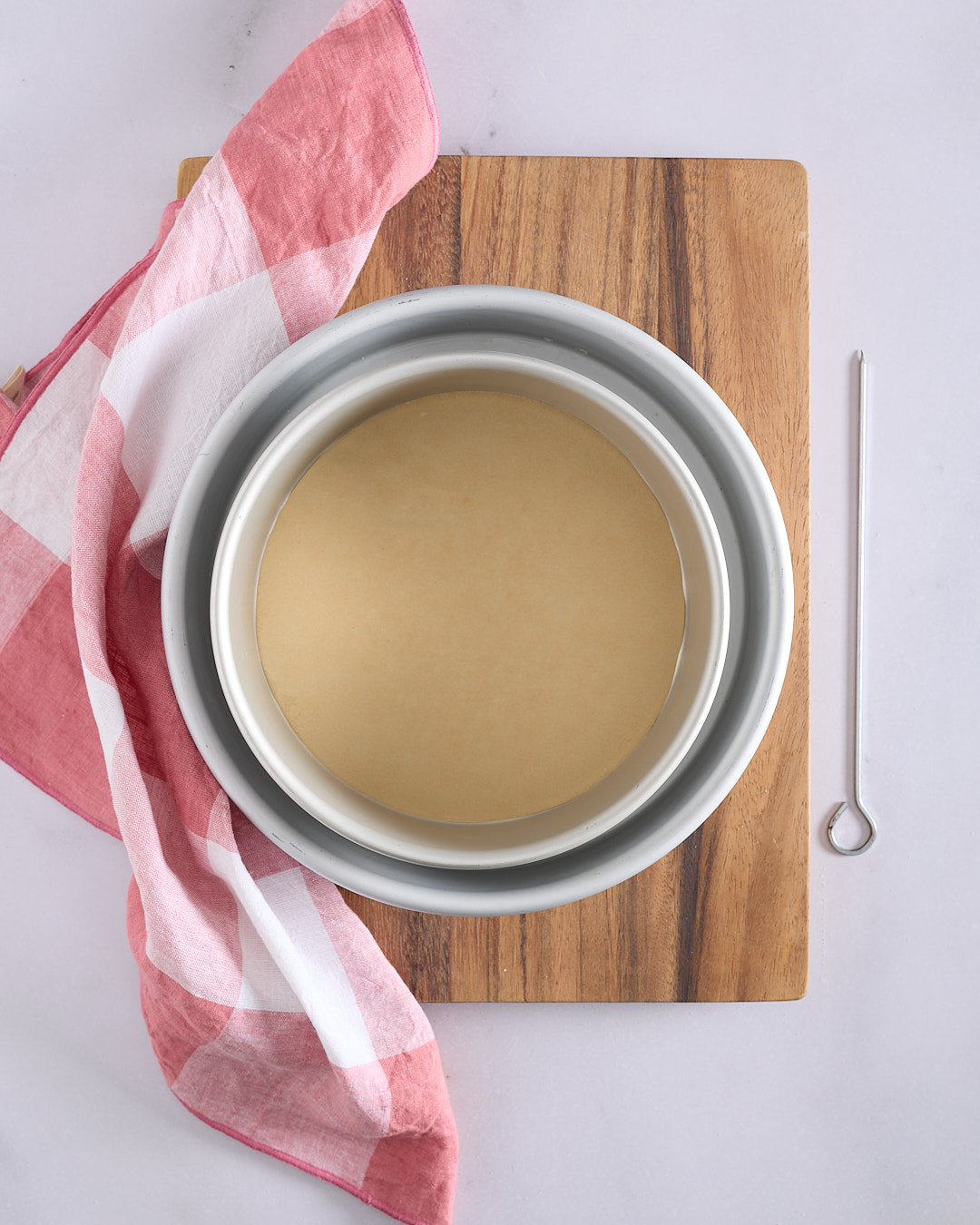 An 8-inch silver cake pan nested inside a 10-inch pan on a wooden board next to a metal skewer and a red and white checkered tea towel.