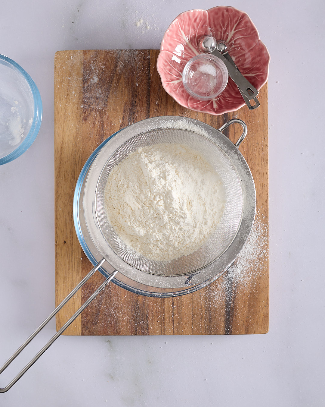 Sifting cake flour through a fine mesh stainless steel sieve into a glass bowl on a wooden board.