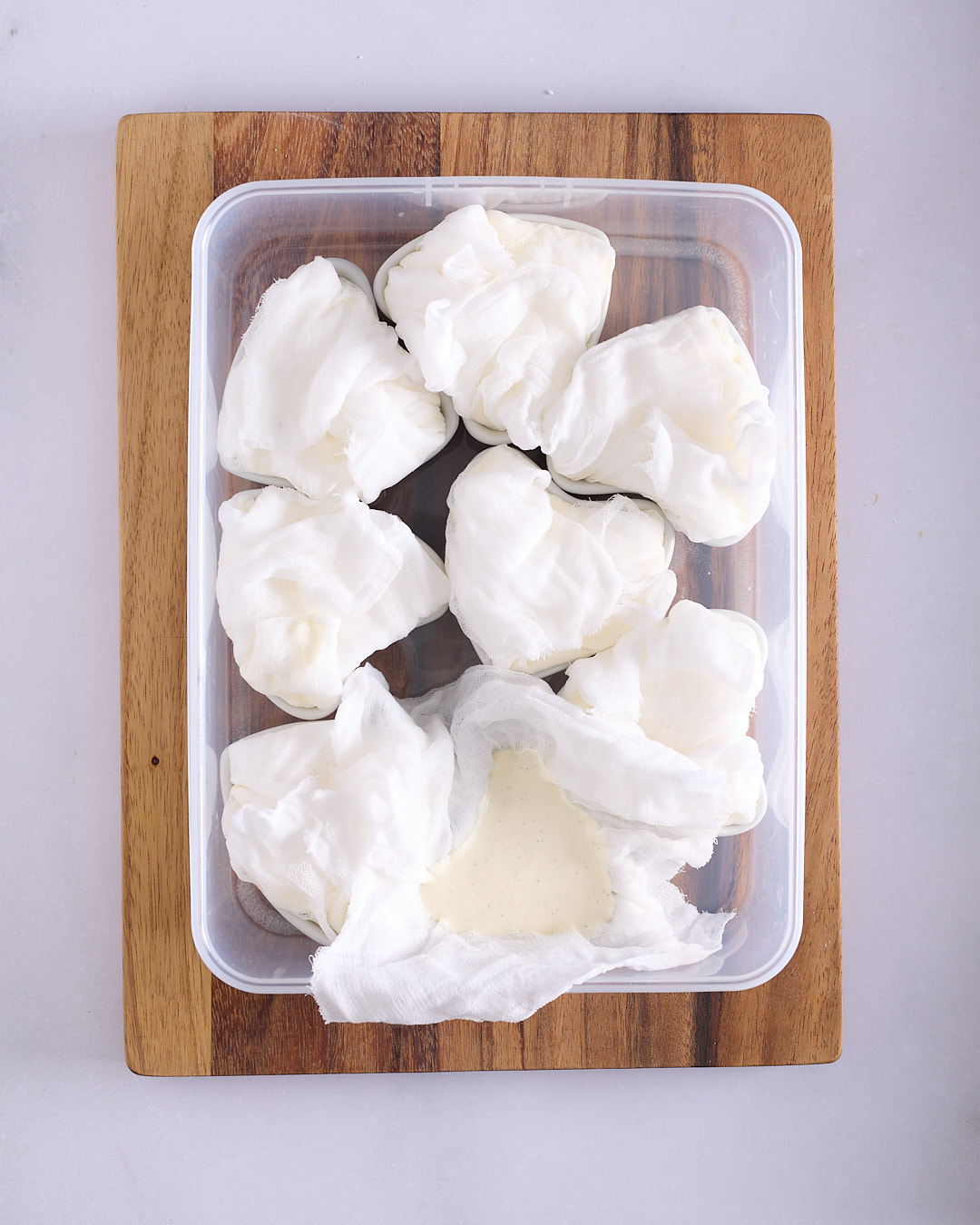 Individual heart shaped moulds filled with yoghurt coeur à la crème and encased in white cheesecloth sitting in a clear plastic container on a wooden board with one mould open to show the vanilla seeds in the mixture.