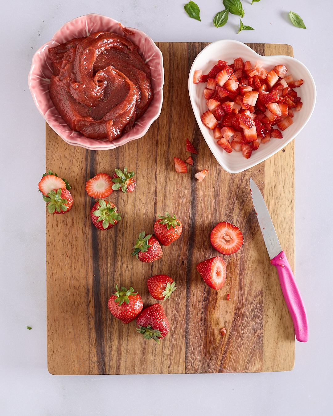 Strawberry mixture in a pink bowl next to finely chopped fresh strawberries in a heart-shaped dish on a wooden board.