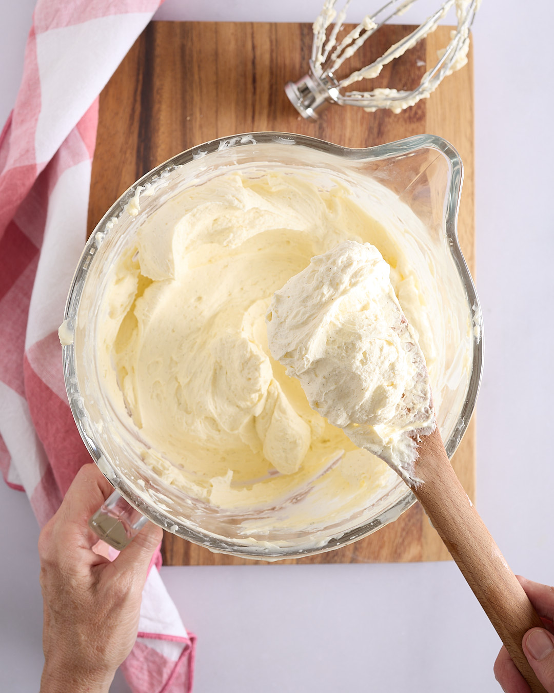 Sandra holding a wooden spoon lifting a large dollop of thick, pale yellow whipped white chocolate ganache over a glass mixing bowl.