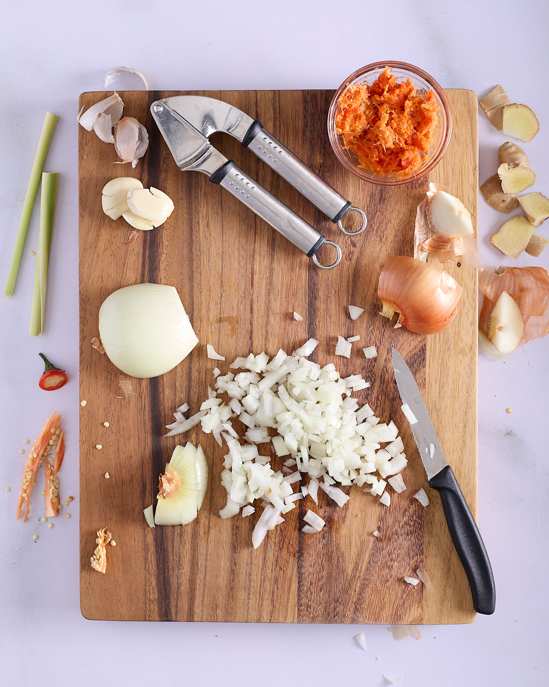 Finely diced brown onion, whole garlic cloves, lemongrass stalks, grated ginger, and fresh chilli on a wooden chopping board showing the preparation of Vietnamese-style pork and chicken meatloaf aromatics, with a garlic press and paring knife alongside.
