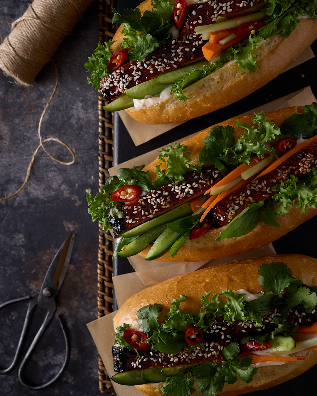 Three vietnamese-style pork and chicken meatloaf bánh mì in mini baguettes on baking paper, loaded with sriracha palm sugar glazed meatloaf slices, fresh cucumber, pickled carrot and celery, red chilli, coriander and mayo, served on a dark moody surface with twine and vintage scissors.