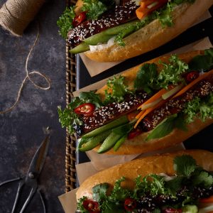 Three vietnamese-style pork and chicken meatloaf bánh mì in mini baguettes on baking paper, loaded with sriracha palm sugar glazed meatloaf slices, fresh cucumber, pickled carrot and celery, red chilli, coriander and mayo, served on a dark moody surface with twine and vintage scissors.