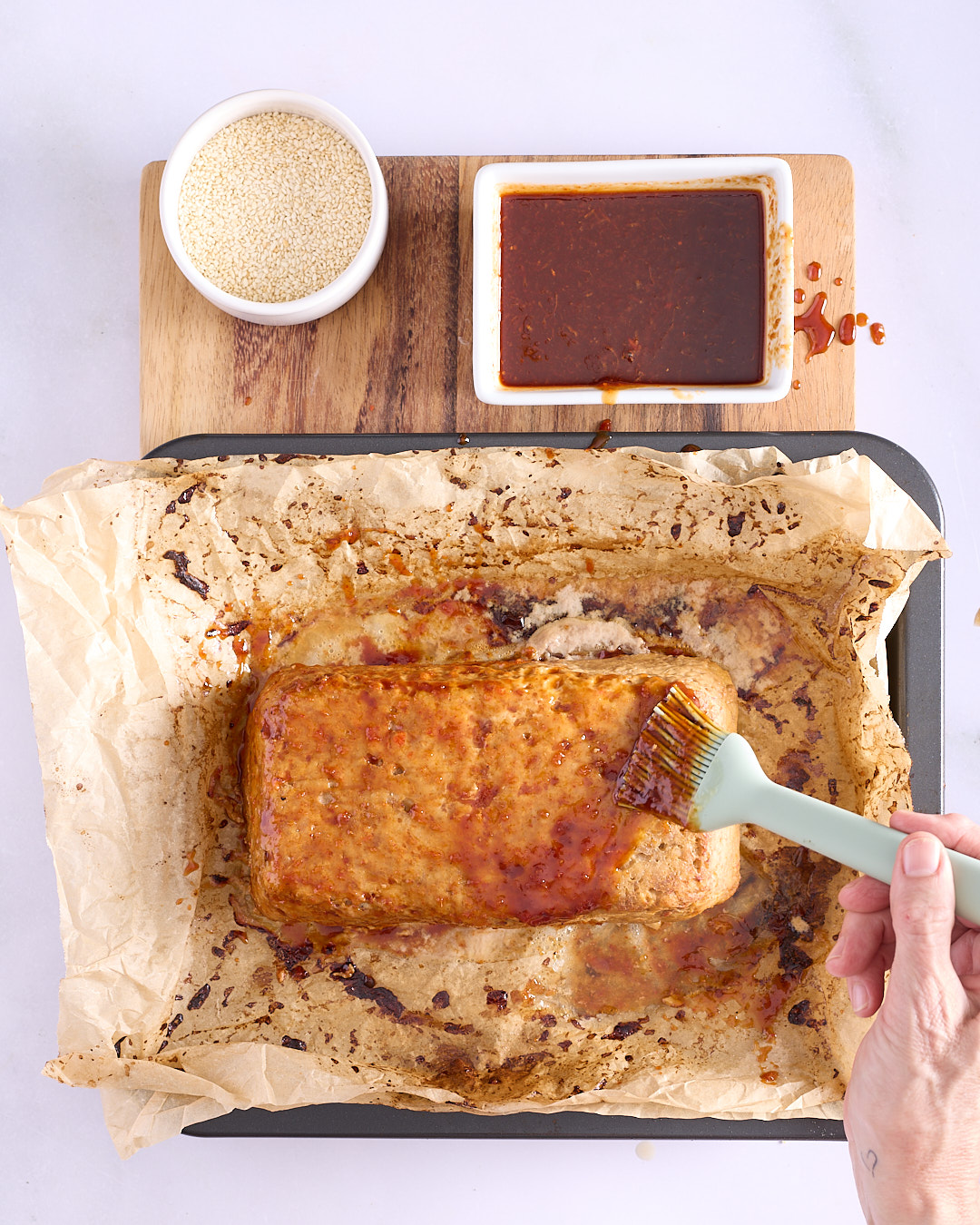 Vietnamese-style pork and chicken meatloaf being basted with sriracha palm sugar glaze on a baking paper lined tray, with toasted sesame seeds and remaining glaze alongside.