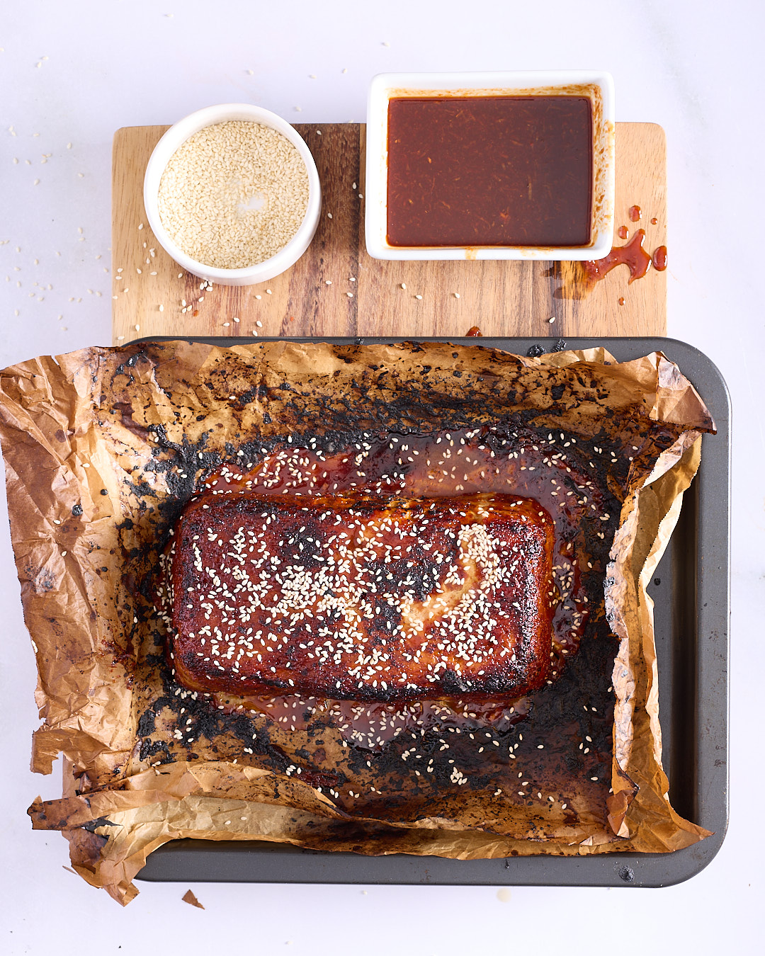 Deeply glazed Vietnamese-style pork and chicken meatloaf topped with toasted sesame seeds on a baking paper lined tray, showing the lacquered finish of the sriracha palm sugar glaze.