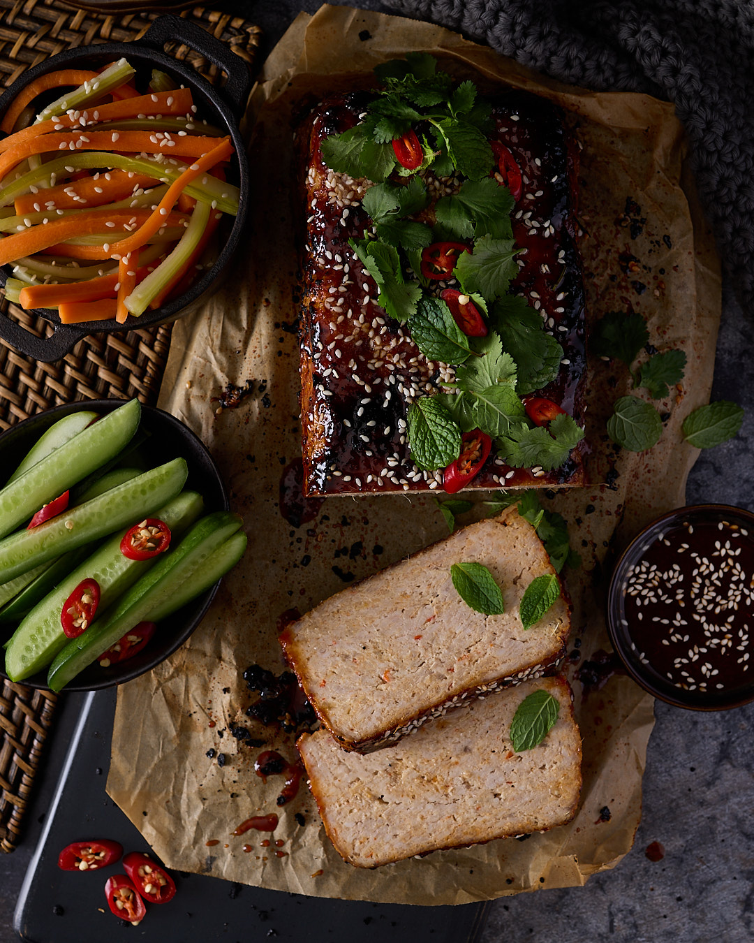 Vietnamese-style pork and chicken meatloaf with sriracha palm sugar glaze and toasted sesame seeds on baking paper, sliced to show the tender interior, served with fresh cucumber batons, quick-pickled carrot and celery, red chilli, coriander and mint.