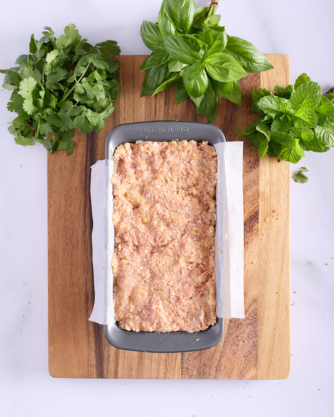 Raw Vietnamese-style pork and chicken meatloaf mixture packed into a lined non-stick loaf pan with a baking paper sling, surrounded by fresh coriander, basil and mint on a wooden board.