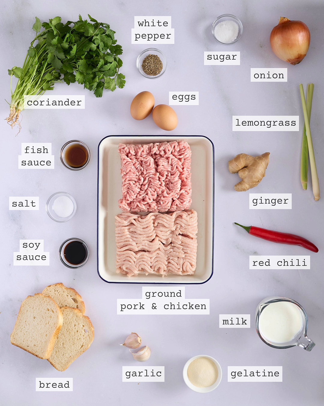Flat lay of ingredients for vietnamese-style pork and chicken meatloaf including ground pork and chicken mince, fish sauce, soy sauce, gelatine, milk, bread, lemongrass, ginger, garlic, red chilli, coriander, eggs, onion, white pepper and sugar, each clearly labelled on a white marble surface.