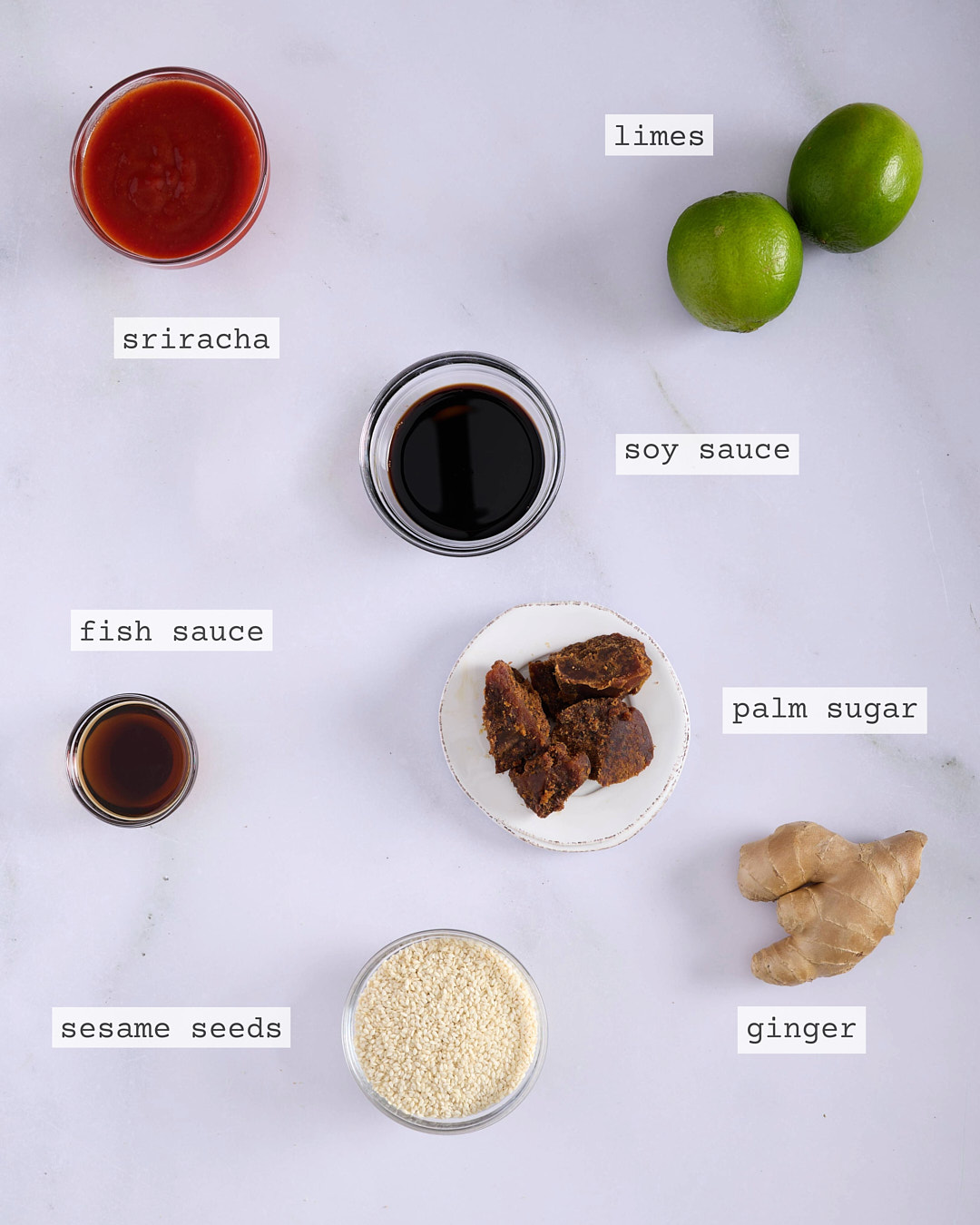 Flat lay of ingredients for Vietnamese-style pork and chicken meatloaf sriracha palm sugar glaze including sriracha, soy sauce, fish sauce, palm sugar, limes, ginger and sesame seeds, each clearly labelled on a white marble surface.