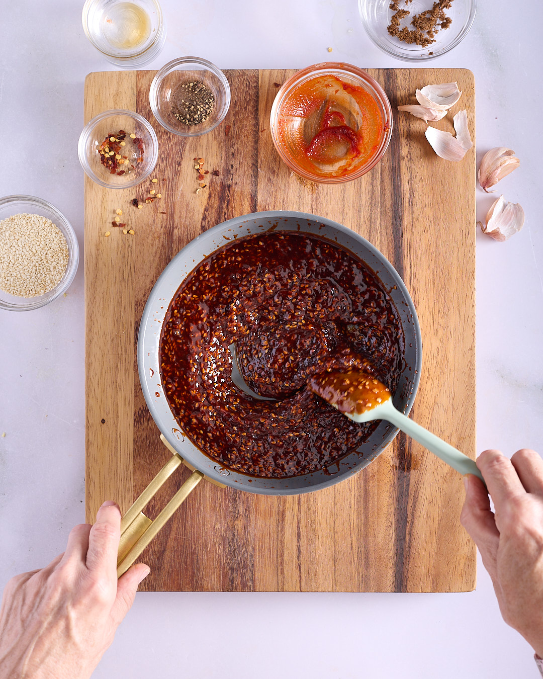 Sandra stirring gochujang BBQ sauce in a small saucepan on a wooden board, with gochujang, chilli flakes, black pepper, sesame seeds and garlic alongside, showing the sauce preparation step for the gochujang pork burger.